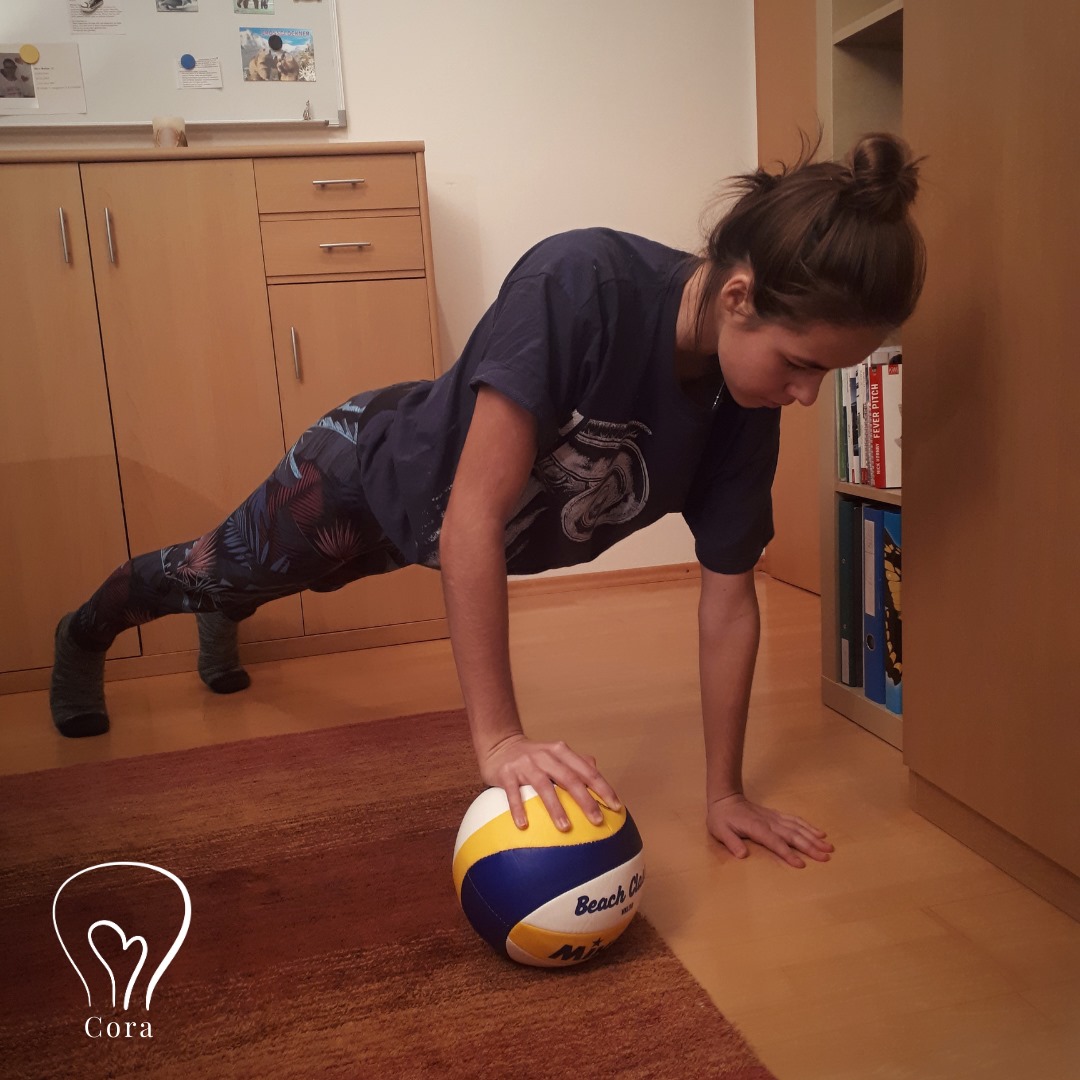 Mädchen in Sportgewand. Gerade macht sie Liegestütz, wobei sie zwischen ihrer rechten Hand und dem Boden einen Volleyball hat.